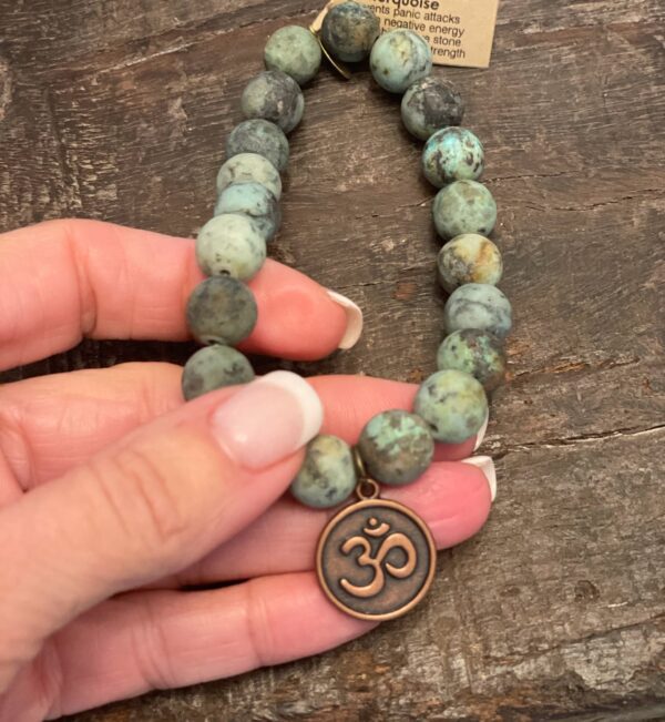 Hand holding a Turquoise Bracelet featuring 10mm matte African turquoise beads and vintage brass om charm.